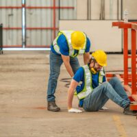 A worker in a hard hat is injured and being helped by another worker. A worker in a hard hat is injured and being helped by another worker.